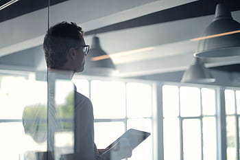 A businessman holding a tablet in an office, looking thoughtfully through a glass wall. HD wallpaper