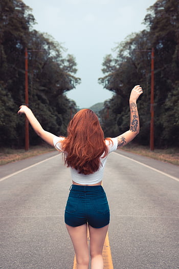 Woman with red hair walking ly down a tree-lined road, showcasing tattoos and casual fashion. HD phone wallpaper