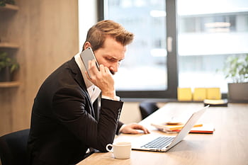 A businessman in a suit making a call while working on a laptop in a modern office setting. HD wallpaper