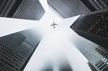 Airplane soaring above fog-enshrouded skyscrapers in an urban setting. HD wallpaper
