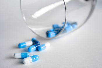 A close-up of blue and white capsules spilling from a glass on a light background. HD wallpaper