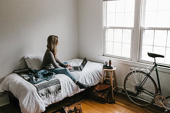 A young woman works on a laptop in a cozy, minimalist bedroom setting, showcasing remote work lifestyle. HD wallpaper