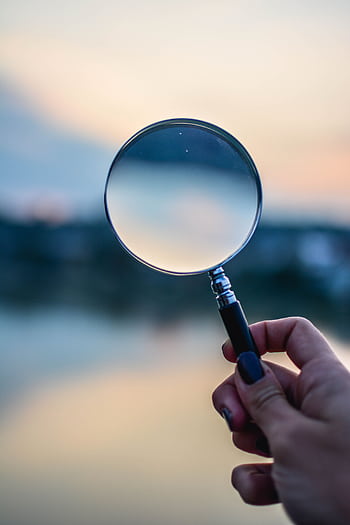 A magnifying glass held by a hand outdoors at sunset, focusing on exploration. HD phone wallpaper