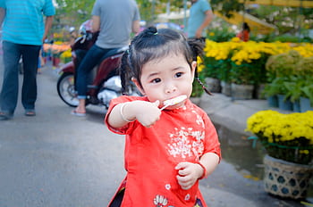 Cute toddler girl in red traditional dress eating a lollipop in a vibrant park setting. HD wallpaper