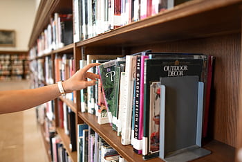 A close-up of a person's hand reaching for a book in a library shelf, suggesting exploration and learning. HD wallpaper