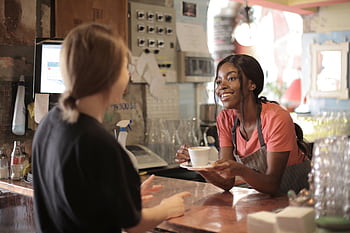 Friendly barista in an apron serves coffee to a customer in a warm, inviting cafe setting. HD wallpaper