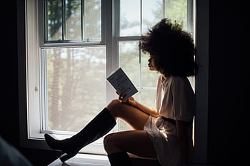 A woman enjoys a peaceful reading session by a window, bathed in natural light. HD wallpaper