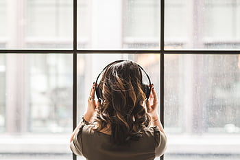 A young woman enjoys music through heads while looking out a large window. HD wallpaper
