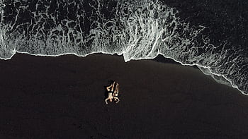 Aerial view of a couple embracing on a black sand beach with waves crashing at Karangasem, Bali. HD wallpaper