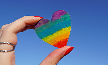 A hand holding a rainbow-colored heart against a clear blue sky, symbolizing love and pride. HD wallpaper