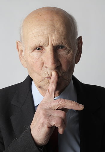 Portrait of a senior man wearing a suit, gesturing for silence in a studio setting. HD phone wallpaper