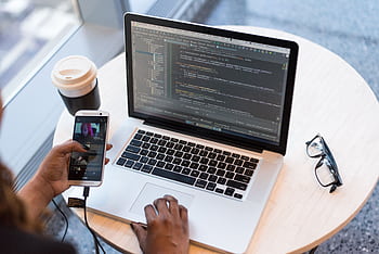 A programmer manages code on a laptop while multitasking with a smart, illustrating modern tech workspace. HD wallpaper