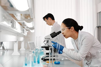 Two scientists in a lab examining samples with a microscope. Focus on scientific research and discovery. HD wallpaper