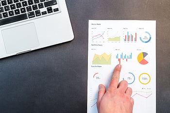 A hand pointing at colorful charts with a laptop in the background on a desk. HD wallpaper