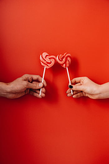Two hands holding heart-shaped lollipops against a vibrant red background, symbolizing love. HD phone wallpaper