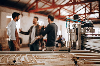 Three men discussing work in a woodworking shop with CNC machinery in focus. HD wallpaper
