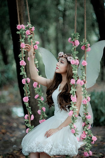A fairy dressed in white, sitting on a flower-adorned swing in a mystical forest setting. HD phone wallpaper