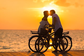 A couple shares a romantic kiss on bicycles during a beautiful sunset at the beach. HD wallpaper