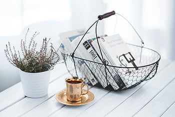 A tranquil home scene featuring a wire basket of books, a golden cup of coffee, and a potted plant on a wooden table. HD wallpaper