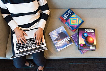 Woman using laptop on sofa, surrounded by programming books, learning coding. HD wallpaper