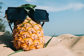 A pineapple with sunglasses sits on a sunny beach, epitomizing summer vacation vibes. HD wallpaper