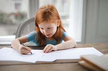 A young girl with red hair focused on drawing at a wooden desk indoors. HD wallpaper