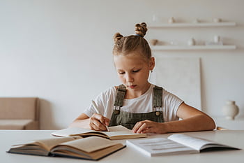 Young girl with braided hair concentrating on her homework at a desk with books, highlighting learning at home. HD wallpaper