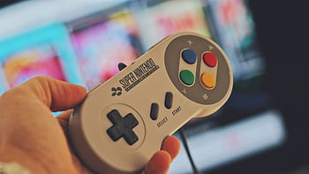 Hand holding a classic Super Nintendo game controller indoors with a blurred screen in the background. HD wallpaper