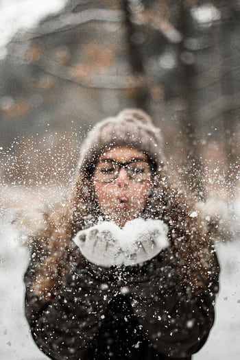 A woman in winter clothing playfully blowing snowflakes outdoors, capturing the joy of a snowy day. HD phone wallpaper