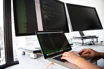 A developer writes code on a laptop in front of multiple monitors in an office setting. HD wallpaper