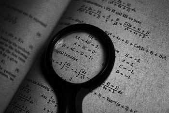 A magnifying glass focuses on mathematics formulas in a book, enhancing study and exploration. HD wallpaper