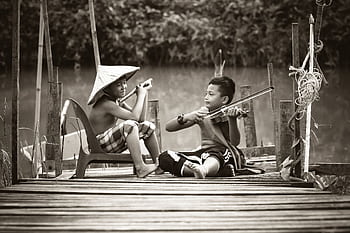 Two children sitting on a dock, playing musical instruments in a serene outdoor setting. HD wallpaper