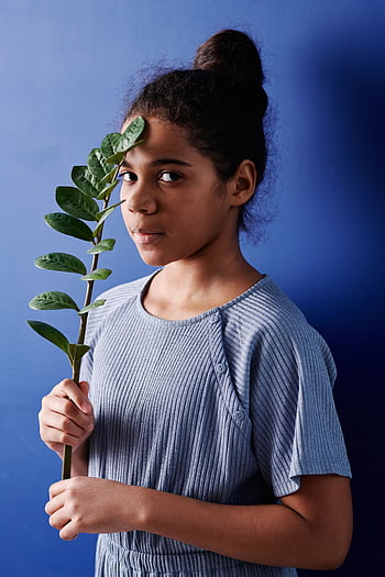 A young girl with a leaf in front of her face stands against a blue backdrop, displaying an artistic vibe. HD phone wallpaper